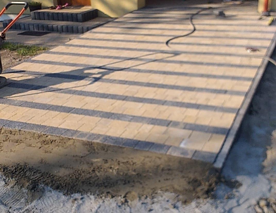 Fragment podjazdu z kostki brukowej w trakcie układania, widoczne przygotowane podłoże i częściowo ułożona nawierzchnia z widocznymi cieniami.