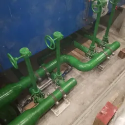 Zestaw zielonych rur z zaworami i pokrętłami, zamontowany przy niebieskiej ścianie, widoczne elementy mocujące i fragment betonowej podłogi.