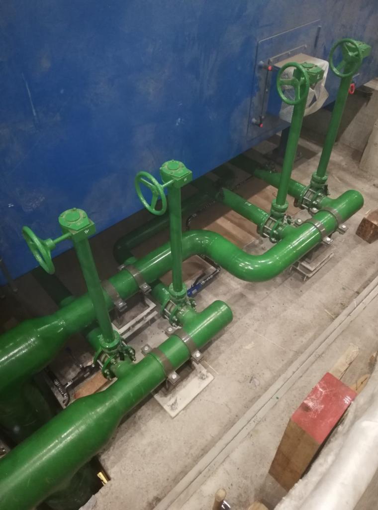 Zestaw zielonych rur z zaworami i pokrętłami, zamontowany przy niebieskiej ścianie, widoczne elementy mocujące i fragment betonowej podłogi.