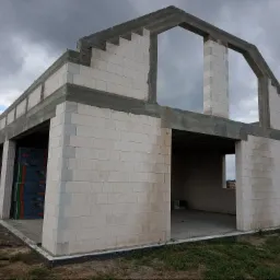 Surowy stan domu z betonu komórkowego w trakcie budowy, widoczne palety z materiałem budowlanym wewnątrz, pochmurne niebo w tle.