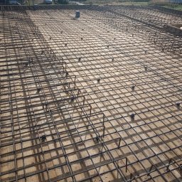 ARTUR SZMALEC Specjalistyczne Usługi Budowalne - Siatka zbrojeniowa z prętów stalowych, ułożona na drewnianym szalunku, przygotowana do zalania betonem, widok z góry, w tle zabudowa jednorodzinna.