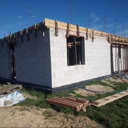 ARTUR SZMALEC Specjalistyczne Usługi Budowalne - Wznoszenie ścian budynku z białych bloczków, widoczne drewniane szalunki i wystające pręty zbrojeniowe, przygotowane do zalewania wieńca, w tle błękitne niebo.