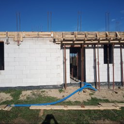 ARTUR SZMALEC Specjalistyczne Usługi Budowalne - Wznoszenie ścian z bloczków białych, widoczne szalunki drewniane i pręty zbrojeniowe, błękitne rury wzdłuż fundamentów, budowa w trakcie realizacji przy słonecznej pogodzie.