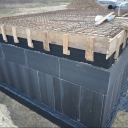 ARTUR SZMALEC Specjalistyczne Usługi Budowalne - Przygotowanie fundamentów: zbrojenie z siatki stalowej na szalunku z desek, izolacja pionowa ścian fundamentowych w kolorze czarnym, widoczny grunt i hałda ziemi w tle.