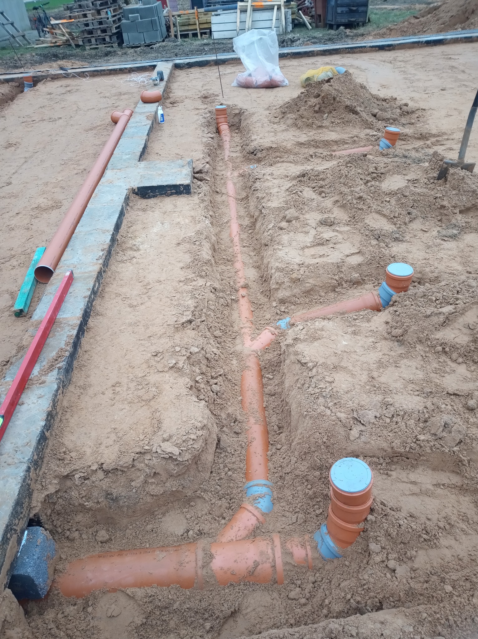 Wykonanie instalacji kanalizacyjnej w gruncie, widoczne rury kanalizacyjne z tworzywa sztucznego w wykopie, poziomica i palety w tle.