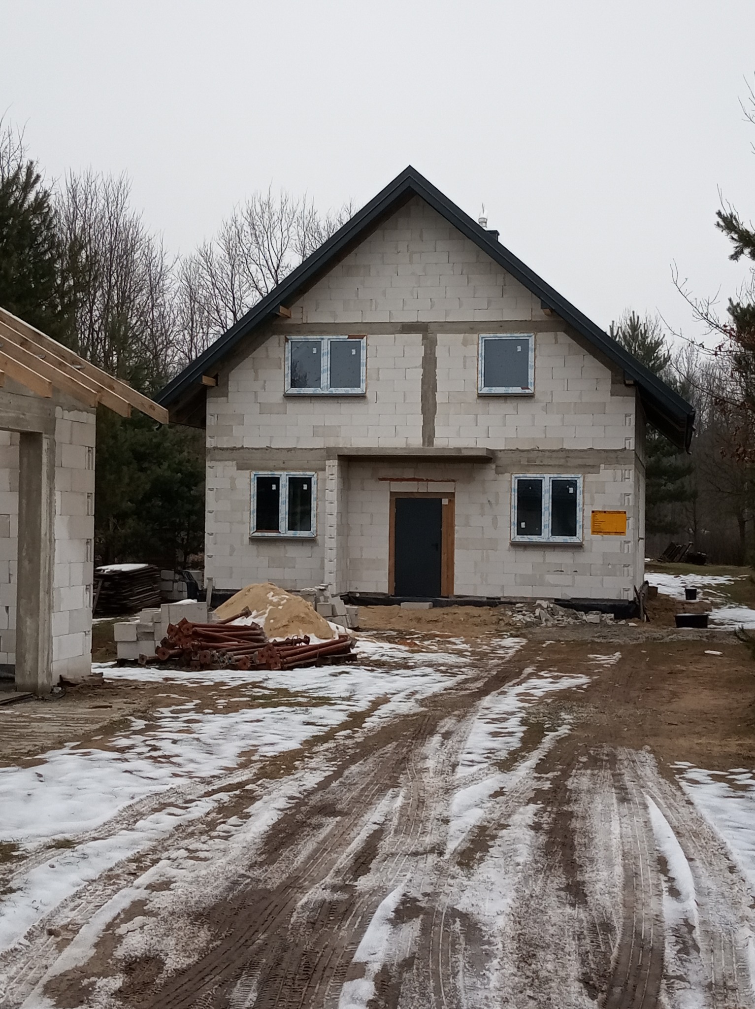 Surowy stan domu z pustaków, z zamontowanymi oknami, widoczne ślady opon w śniegu i piasek na budowie, obok powstający budynek gospodarczy z drewnianą konstrukcją dachu.