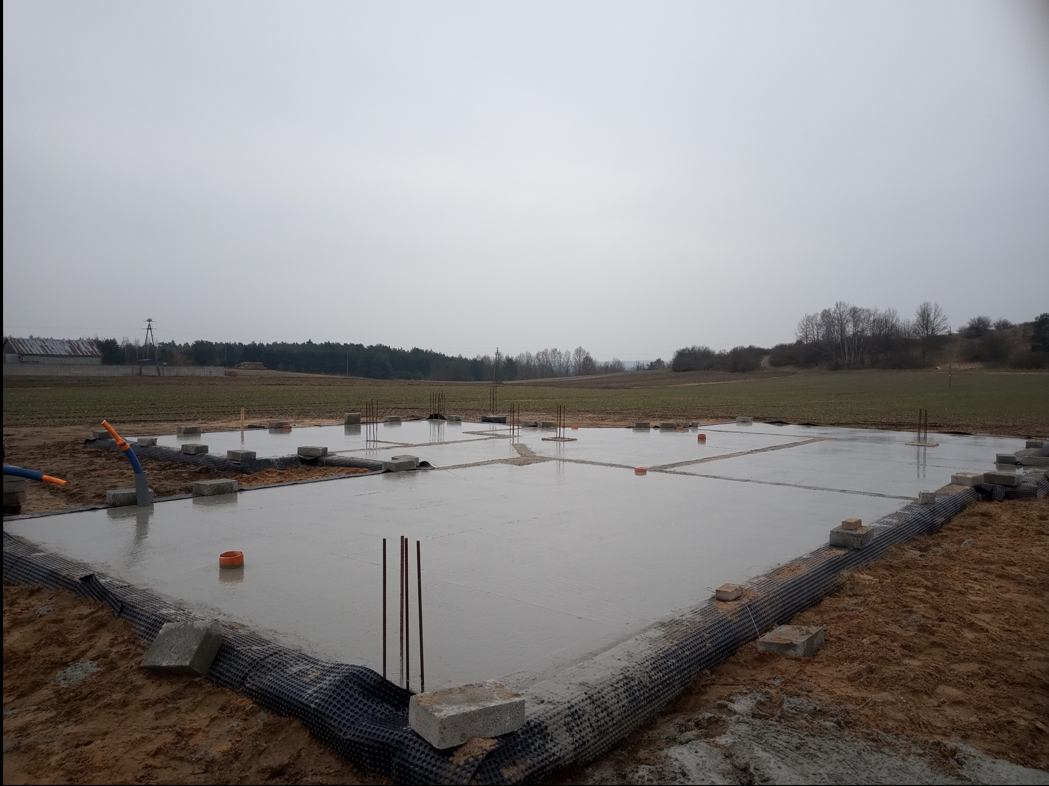 Świeżo wylana płyta fundamentowa z widocznymi prętami zbrojeniowymi, otoczona czarną folią kubełkową i obciążona betonowymi bloczkami, na tle pola i lasu w pochmurny dzień.