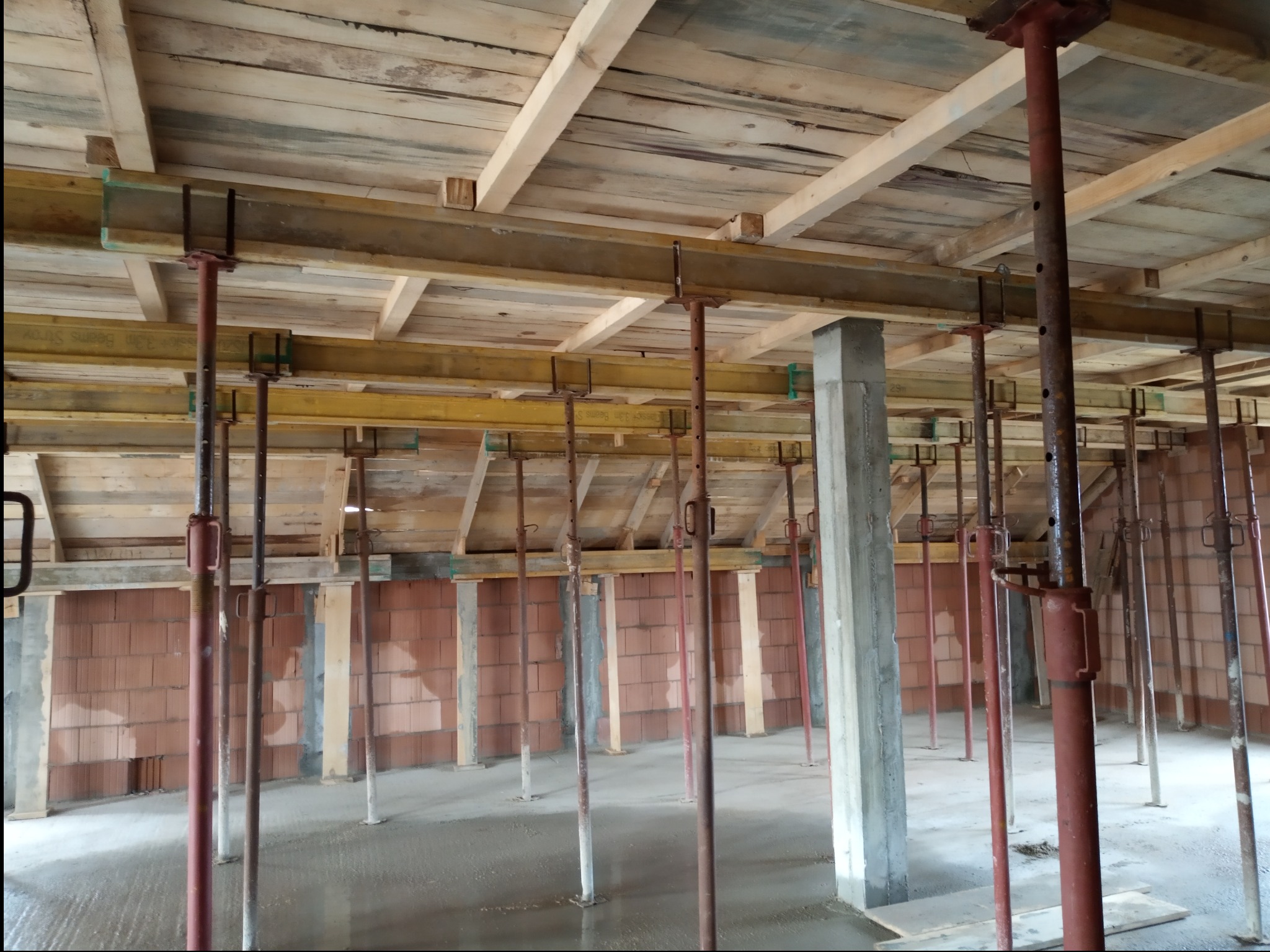 Konstrukcja szalunkowa stropu z podporami teleskopowymi, widoczne ceglane ściany i wylany beton na podłodze.