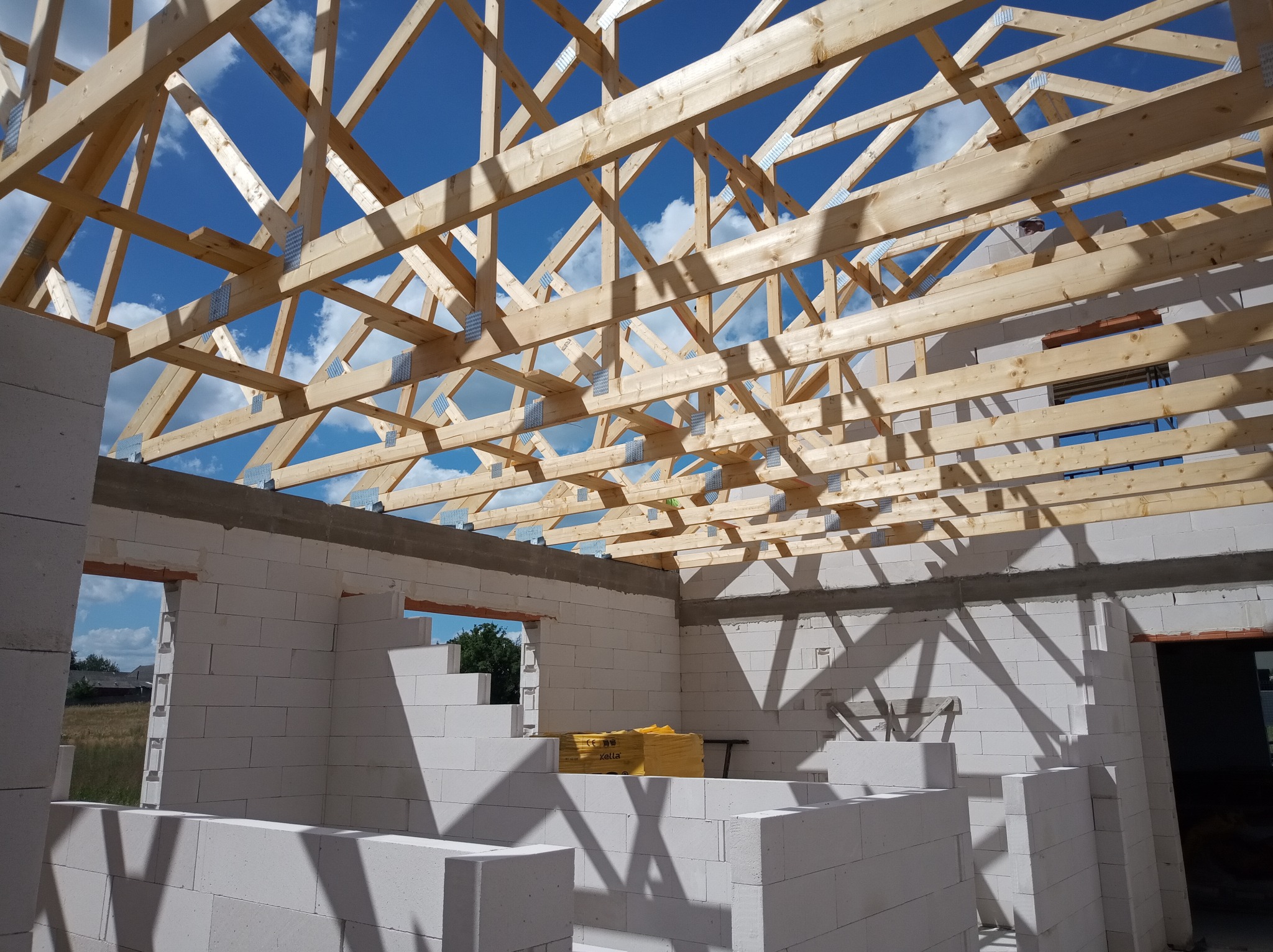 Surowy stan domu murowanego z drewnianą konstrukcją dachu w trakcie budowy, widoczne bloczki z betonu komórkowego i niebo w tle.