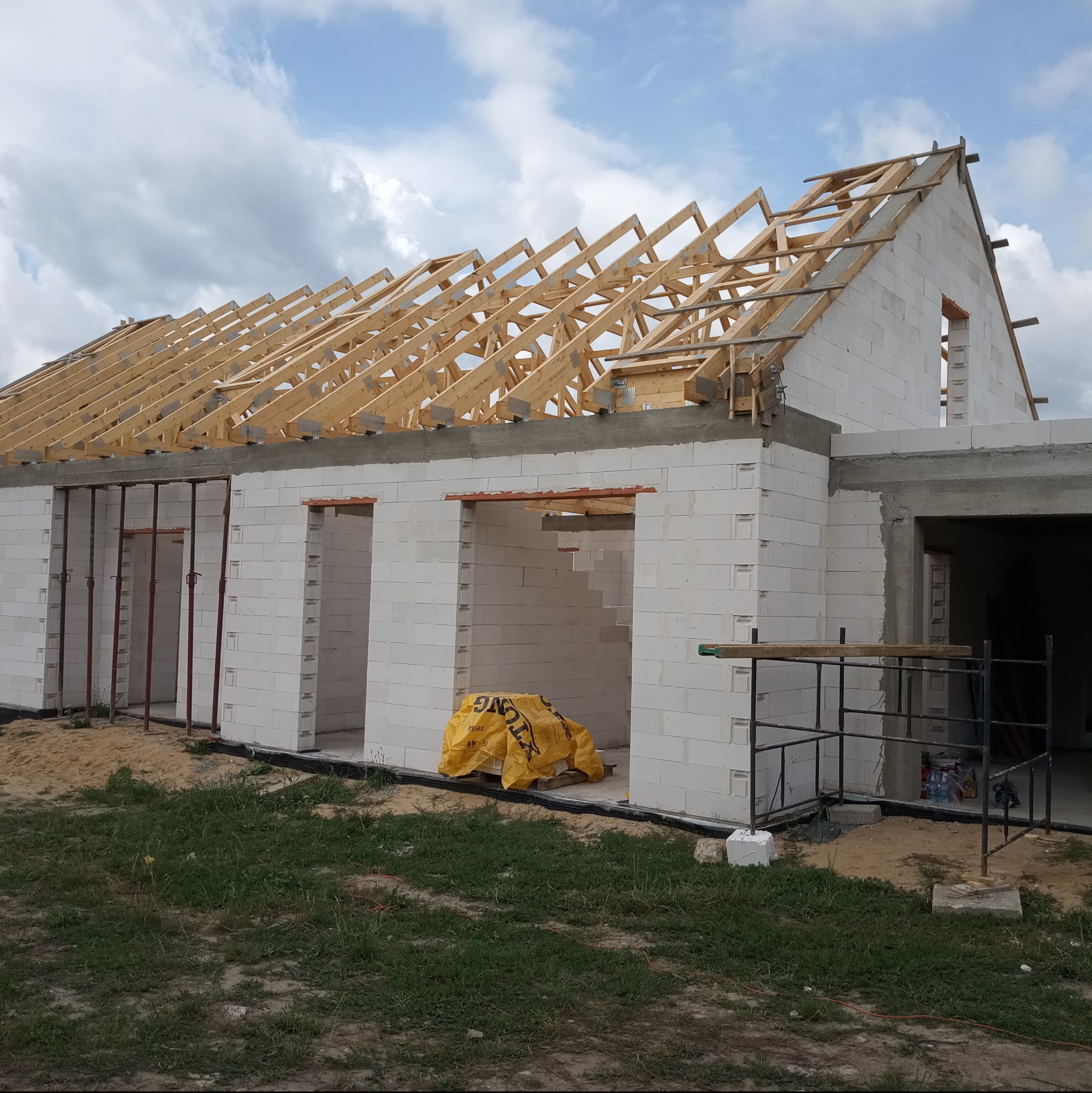 Wznoszenie ścian budynku z bloczków betonu komórkowego; widoczna konstrukcja drewniana dachu, podpory stalowe w otworach okiennych i drzwiowych, oraz rusztowanie.