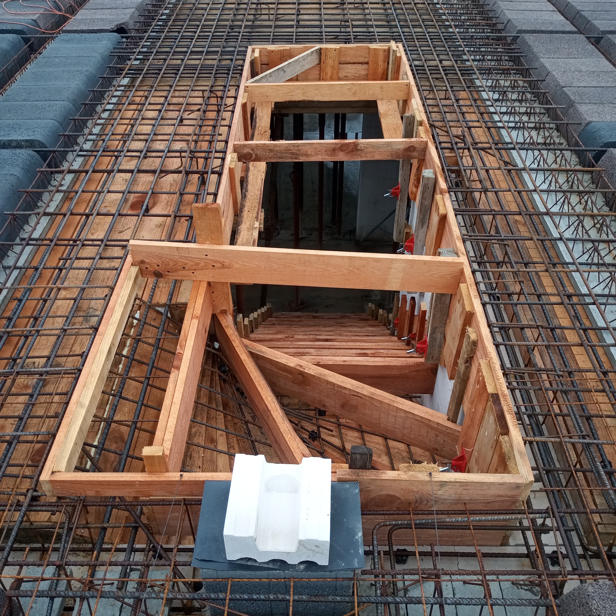 Widok z góry na szalunek z drewna i zbrojenie stalowe przygotowane do zalania betonem, z widocznym otworem w stropie i elementem dystansowym z betonu architektonicznego.