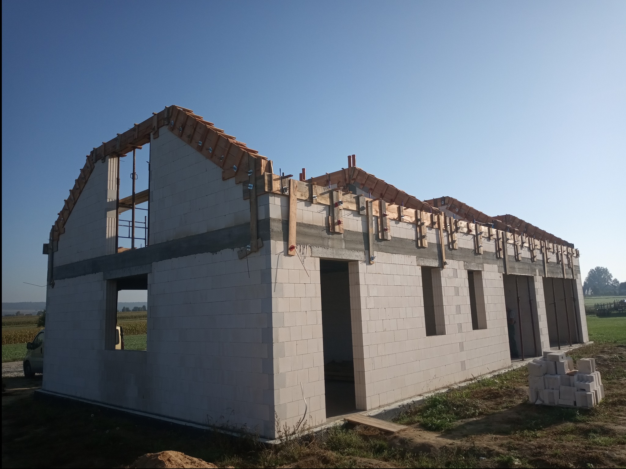 Budowa domu jednorodzinnego w stanie surowym otwartym, ściany z białego bloczka, widoczne drewniane elementy konstrukcji dachu, błękitne niebo w tle.