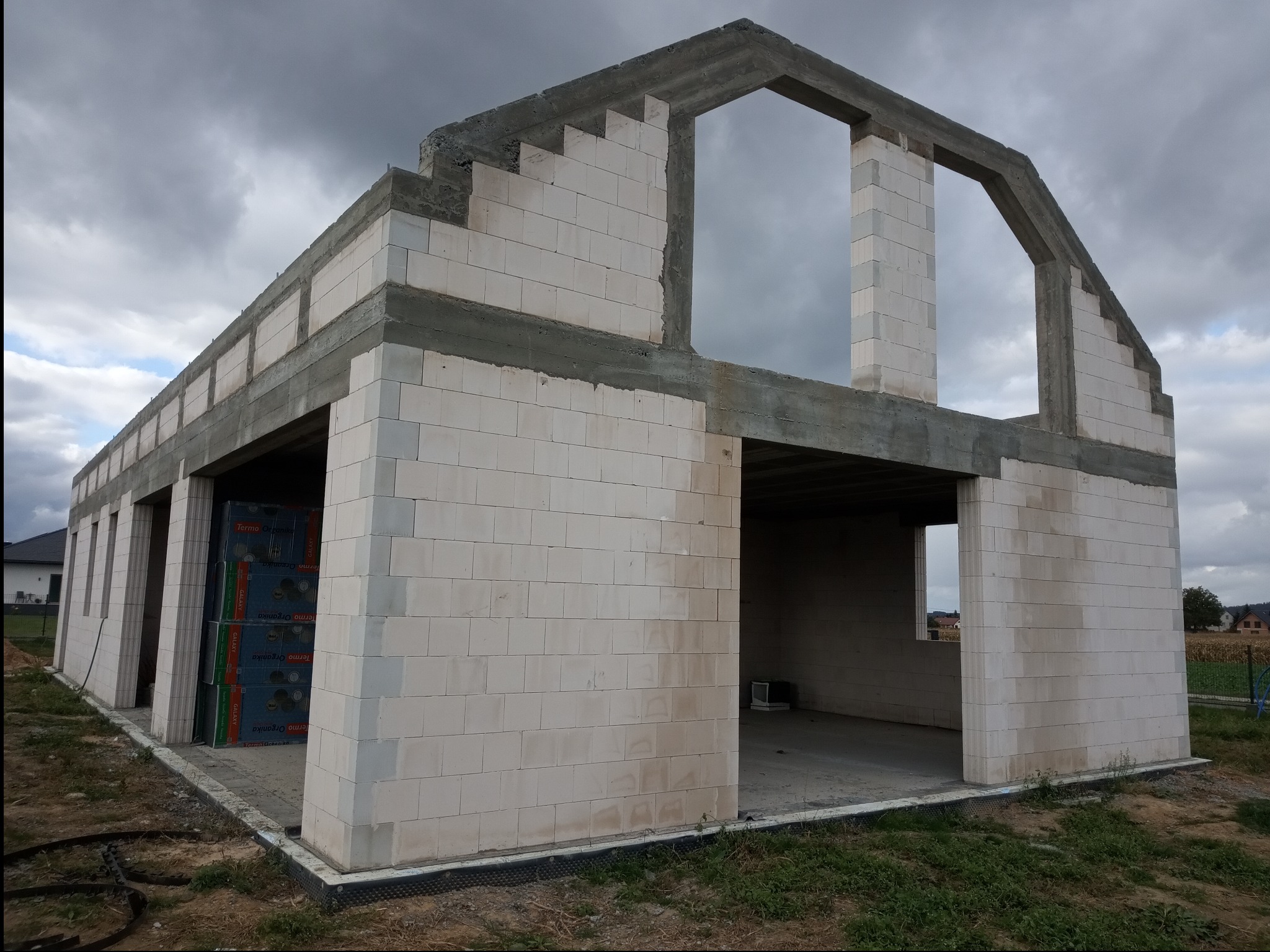 Surowy stan domu z betonu komórkowego w trakcie budowy, widoczne palety z materiałem budowlanym wewnątrz, pochmurne niebo w tle.