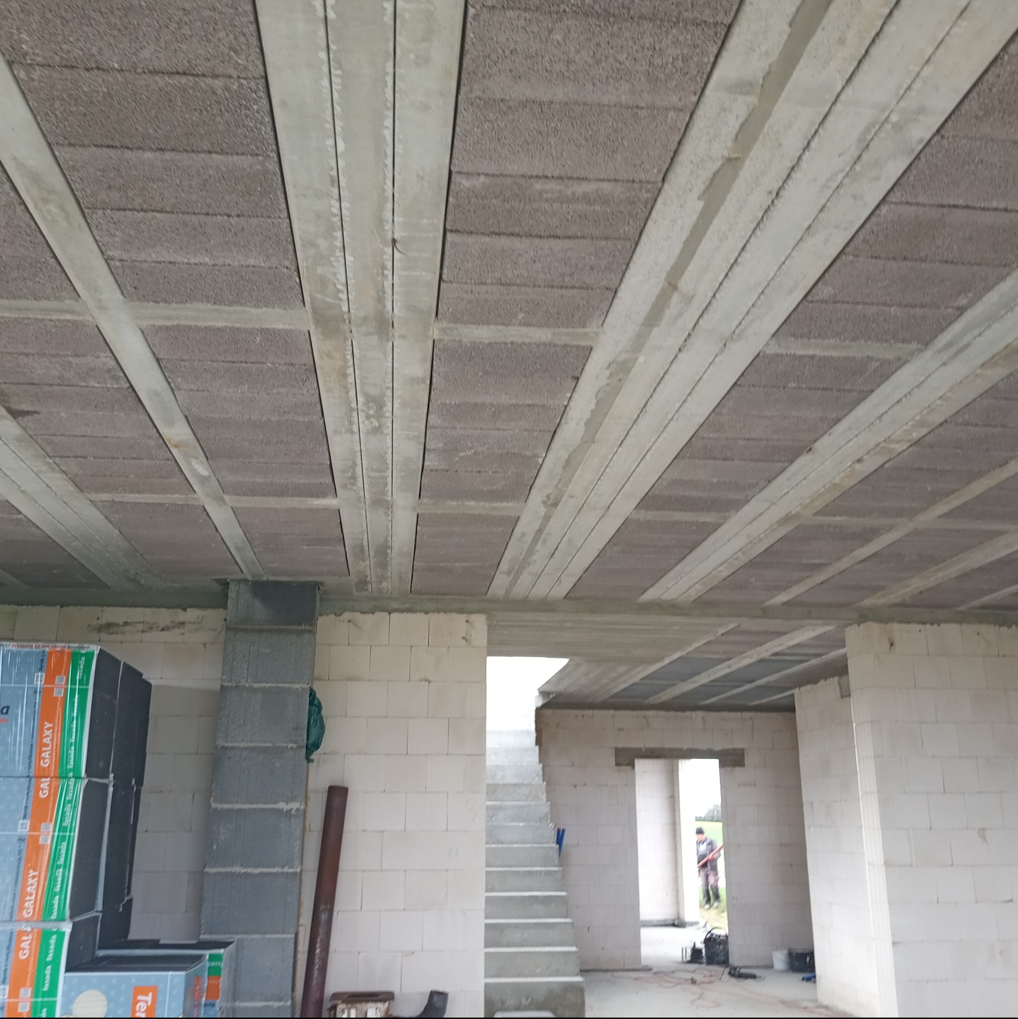 Wnętrze surowego domu w trakcie budowy, widok na strop Teriva z betonowymi belkami i bloczkami, ściany z białych bloczków, w tle schody i otwarte przejścia.