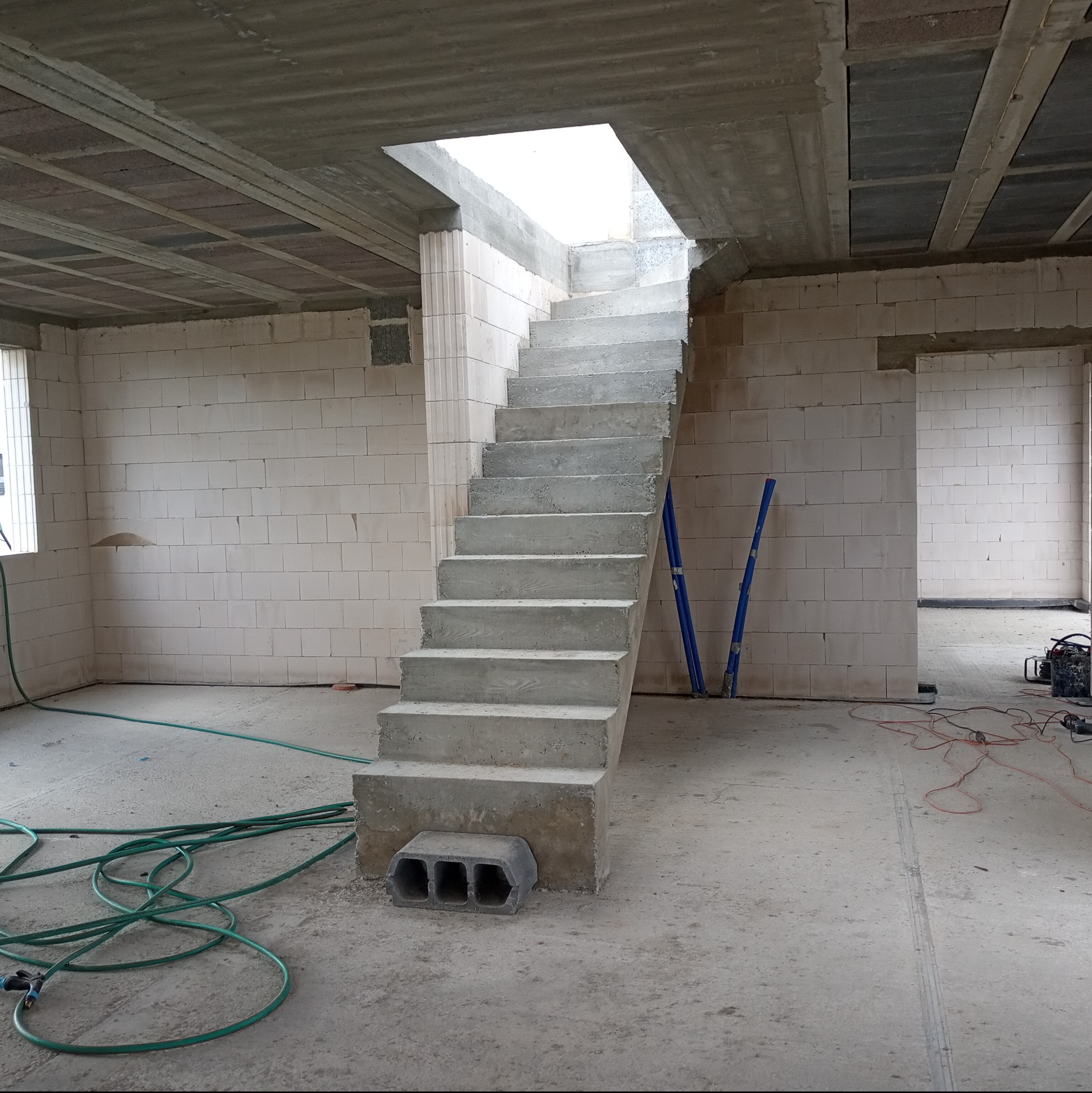 Surowe betonowe schody prowadzące do otworu w stropie wewnątrz budynku z pustaków, widoczne narzędzia i węże na podłodze.