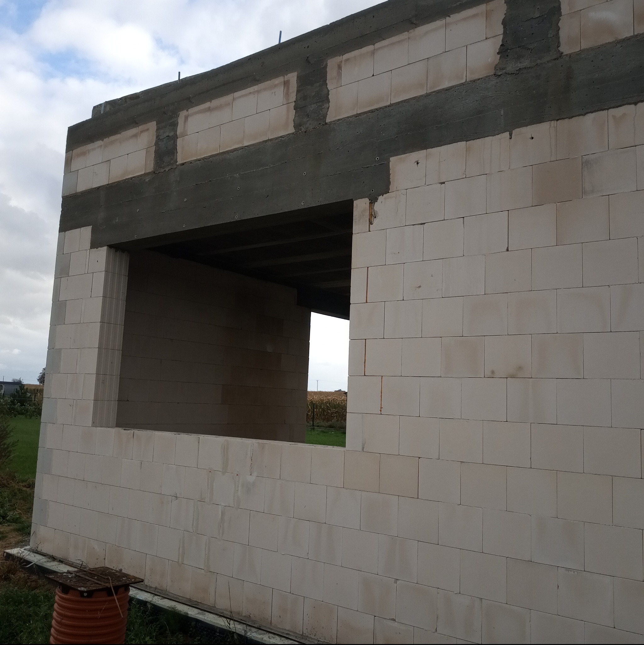 Ściana domu w trakcie budowy z białych bloczków, widoczne nadproże okienne z betonu, w tle pole i pochmurne niebo.
