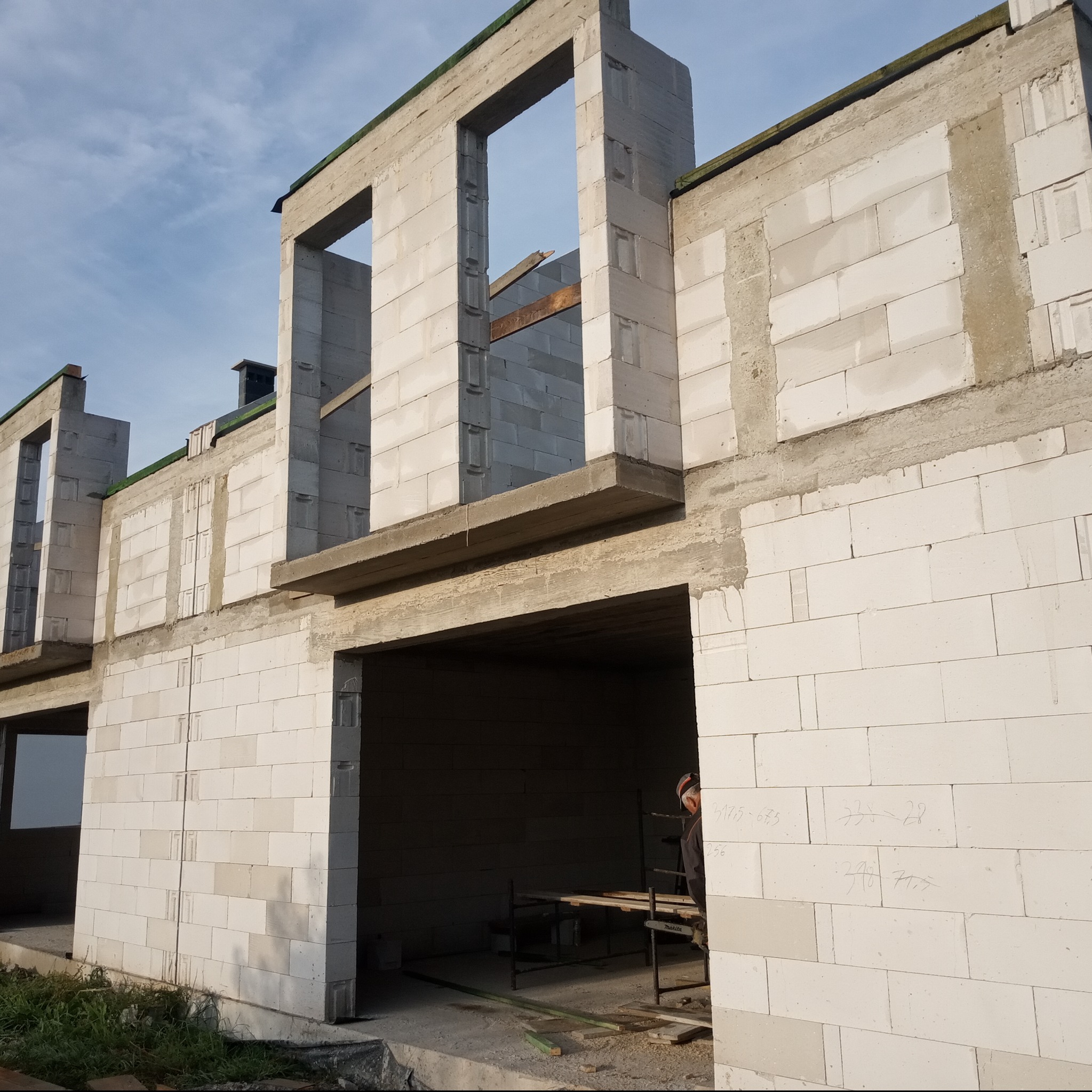 Surowy stan budynku z betonu komórkowego, widoczne otwory okienne i drzwiowe, częściowo otynkowane ściany i zapiski na murze.