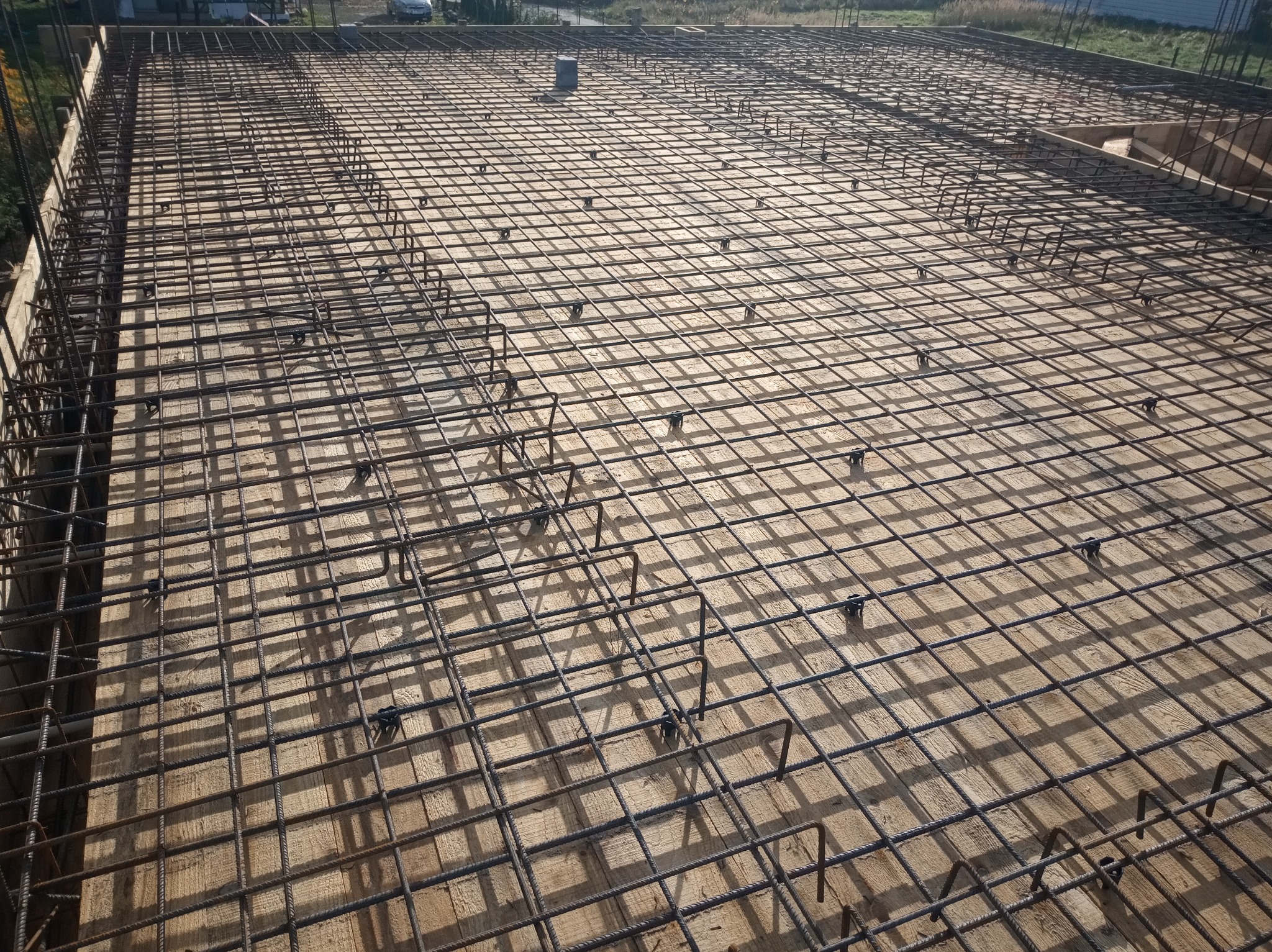Siatka zbrojeniowa z prętów stalowych, ułożona na drewnianym szalunku, przygotowana do zalania betonem, widok z góry, w tle zabudowa jednorodzinna.