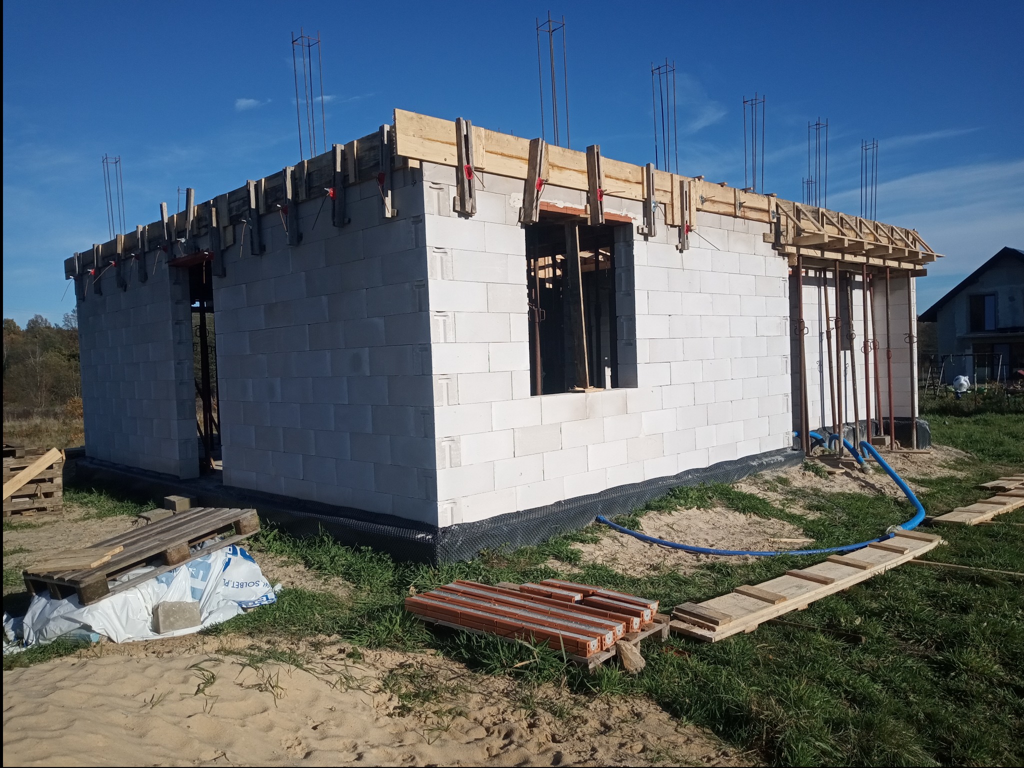 Wznoszenie ścian budynku z białych bloczków, widoczne drewniane szalunki i wystające pręty zbrojeniowe, przygotowane do zalewania wieńca, w tle błękitne niebo.