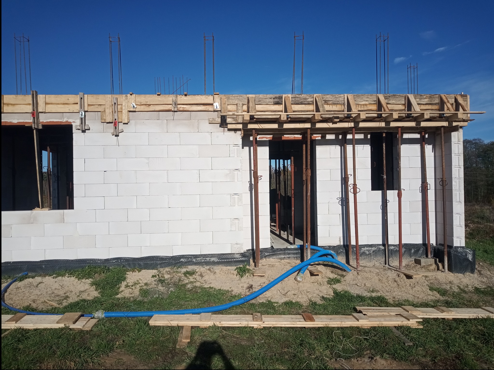 Wznoszenie ścian z bloczków białych, widoczne szalunki drewniane i pręty zbrojeniowe, błękitne rury wzdłuż fundamentów, budowa w trakcie realizacji przy słonecznej pogodzie.