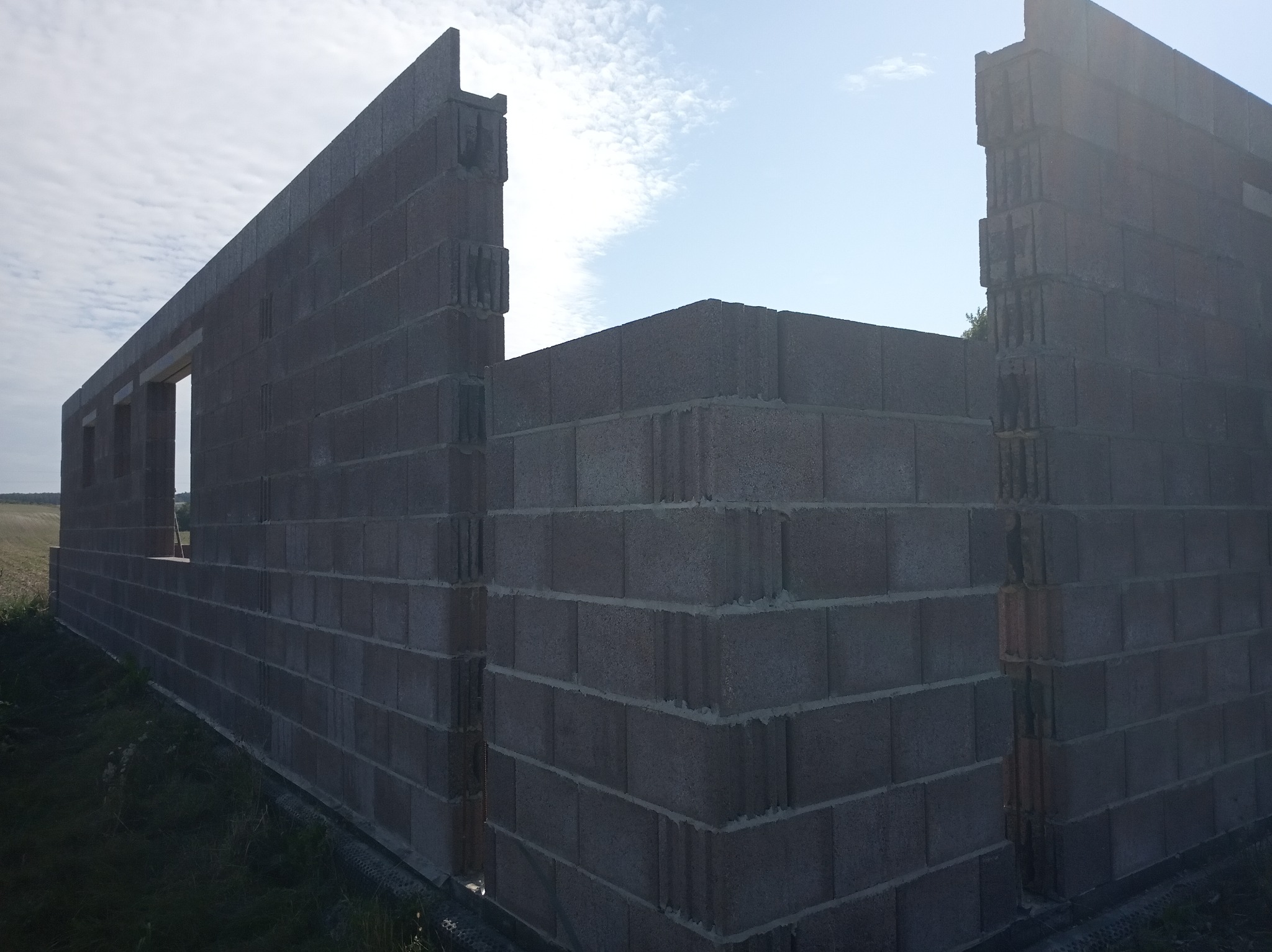 Wznoszone ściany domu z szarego bloczka betonowego, widoczne otwory okienne i narożnik budynku, na zewnątrz pole i błękitne niebo z chmurami.