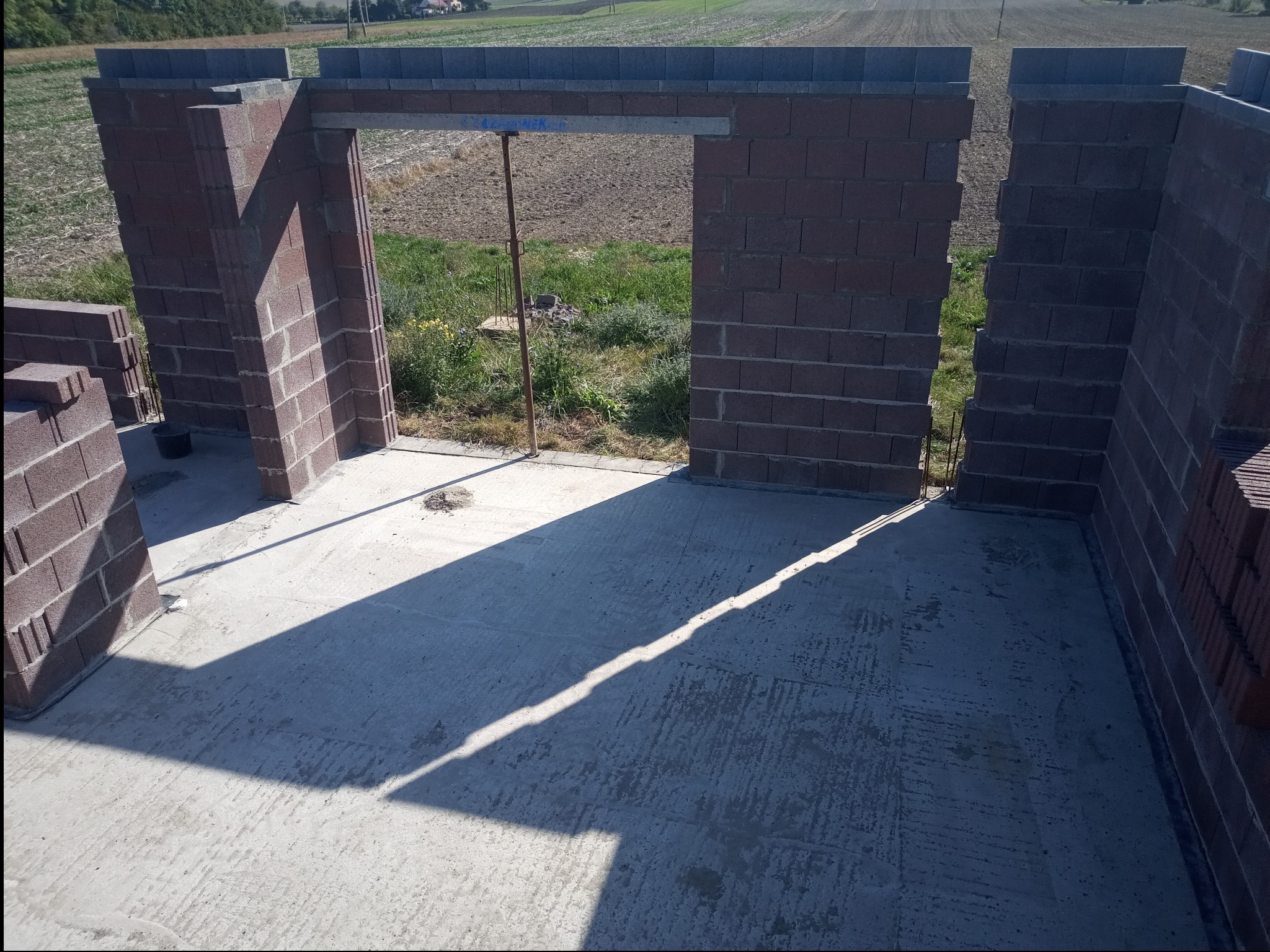 Wznoszone ściany z bloczków na fundamencie, widoczny szalunek nad otworem drzwiowym i zapas bloczków obok fragmentu ściany, w tle pole uprawne.