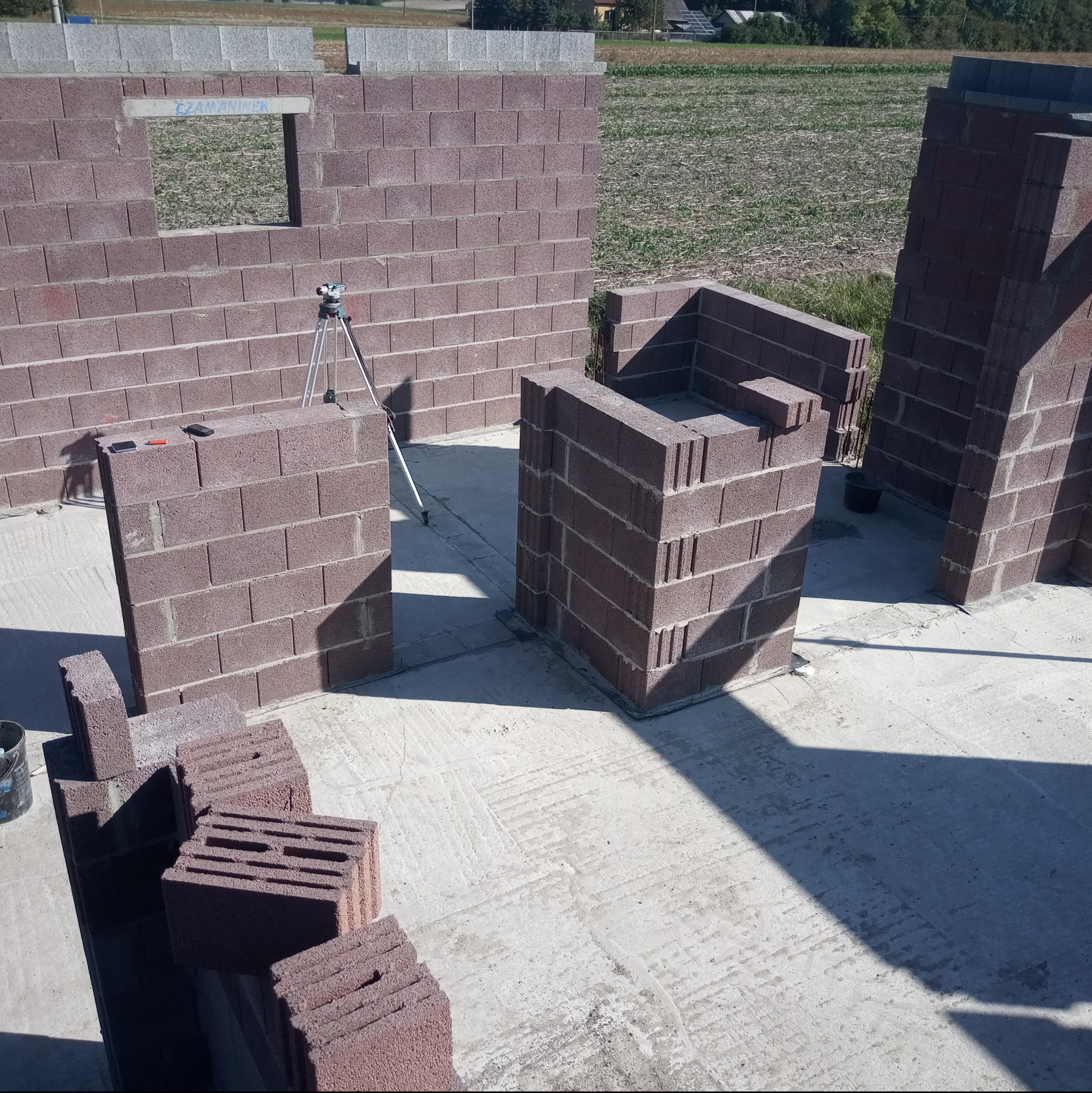 Wznoszenie ścian z bloczków betonu komórkowego, widoczne fragmenty ścian, niwelator optyczny na statywie oraz bloczki czekające na ułożenie.