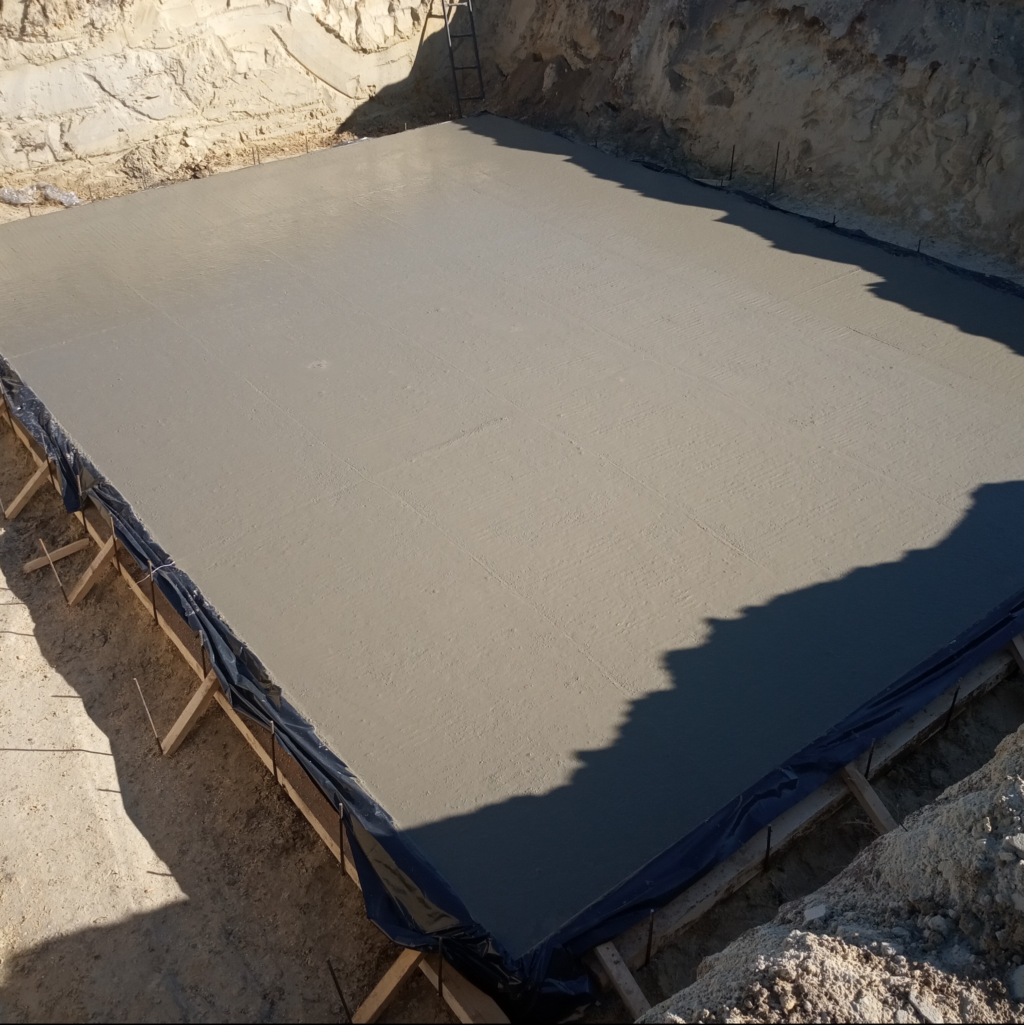 Wylany, świeży beton fundamentowy w wykopie, zabezpieczony folią i drewnianym szalunkiem, w tle widoczna drabina.