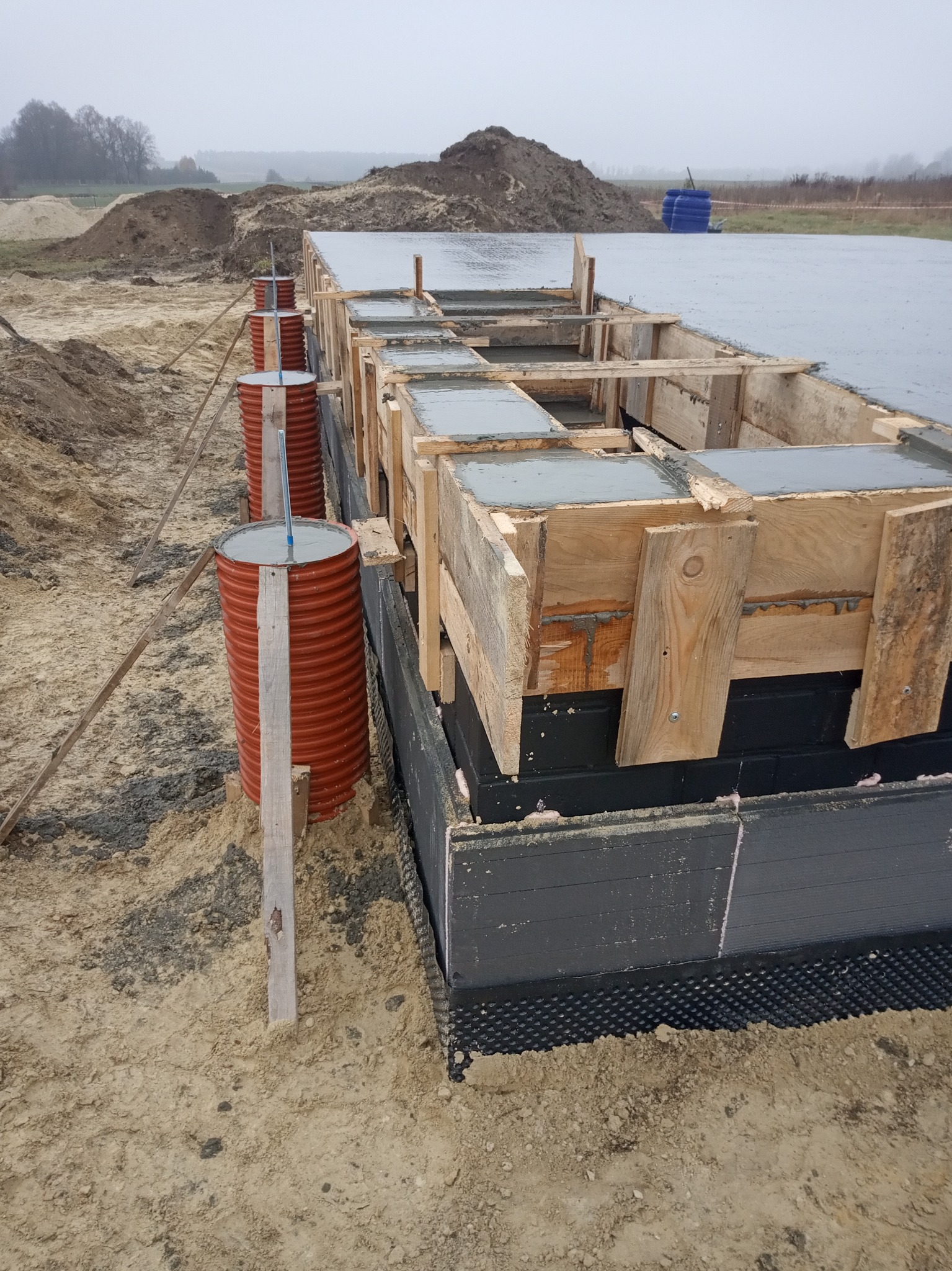 Wylany beton w szalunku fundamentowym z izolacją pionową, wsparty na ziemi za pomocą beczek i drewnianych rozpórek.