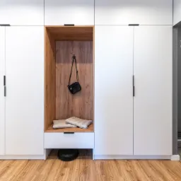 Biała szafa z drewnianą wnęką z poduszkami i torebką, robot sprzątający pod spodem, w tle wieszak na ubrania z napisem 'Wszędzie dobrze, w domu najlepiej'.