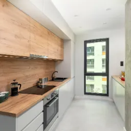 Nowoczesna, wąska kuchnia z drewnianymi frontami szafek, białymi dolnymi szafkami i blatem, z czarną armaturą i ekspres do kawy na blacie, widok na budynek za oknem.