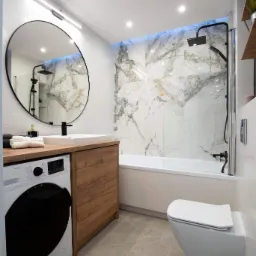 Nowoczesna łazienka z drewnianym blatem i szafką pod umywalką, okrągłym lustrem z czarną ramą, białą pralką, marmurowymi płytkami podświetlonymi na niebiesko, wiszącą toaletą i szarą podłogą...