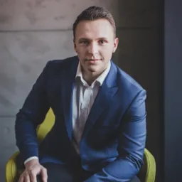 Portret młodego mężczyzny w niebieskim garniturze i białej koszuli, siedzącego na żółtym krześle na tle szarej ściany.