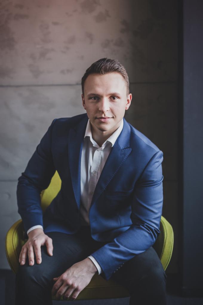 Portret młodego mężczyzny w niebieskim garniturze i białej koszuli, siedzącego na żółtym krześle na tle szarej ściany.