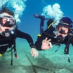 Dw&oacute;ch nurk&oacute;w w pełnym ekwipunku pod wodą, jeden pokazuje znak OK, widoczny piaszczysty grunt i łańcuch.