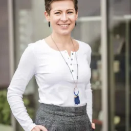 Uśmiechnięta kobieta z krótkimi włosami w białej bluzce i szarych spodniach w kratę, stojąca na tle szklanej witryny.