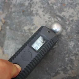 Higrometr z wyświetlaczem wskazującym wilgotność materiału, przykładany do szarej, porowatej powierzchni.