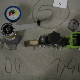 Pomiar wytrzymałości betonu przy użyciu specjalistycznego urządzenia z dwoma wskaźnikami, na powierzchni zaznaczone wyniki pomiarów kredą: 1,00 i 1,40, obok leży ołówek, a na ścianie narysowane...