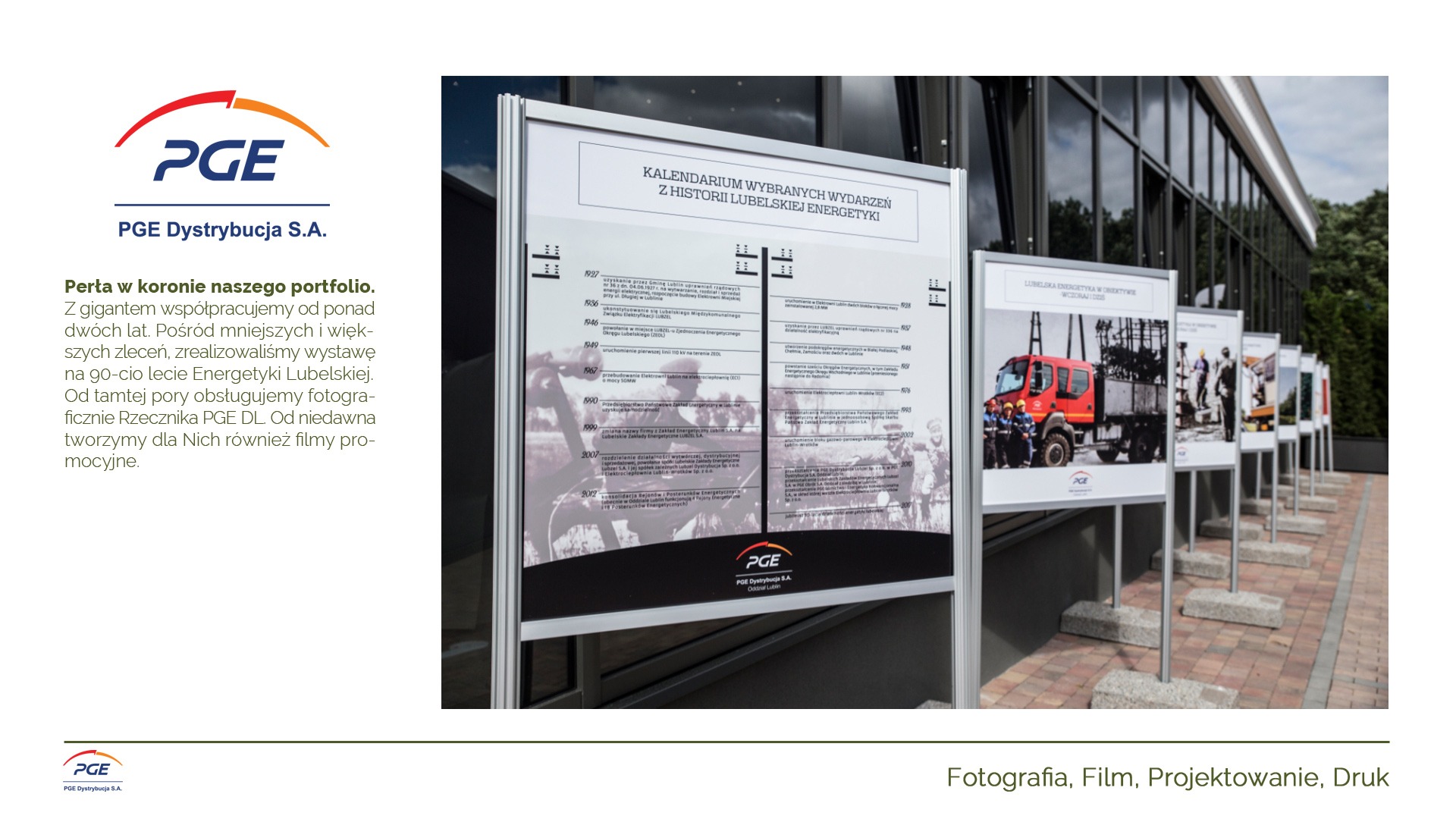 Wystawa plenerowa z planszami prezentującymi historię Lubelskiej Energetyki, z widocznymi zdjęciami i kalendarium wydarzeń, na tle nowoczesnego budynku.