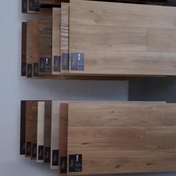 DAIR PHU Alicja Durmała - Ekspozycja próbek podłogowych paneli i parkietów w różnych odcieniach drewna, z etykietami producenta, umieszczonych na półkach w sklepie.