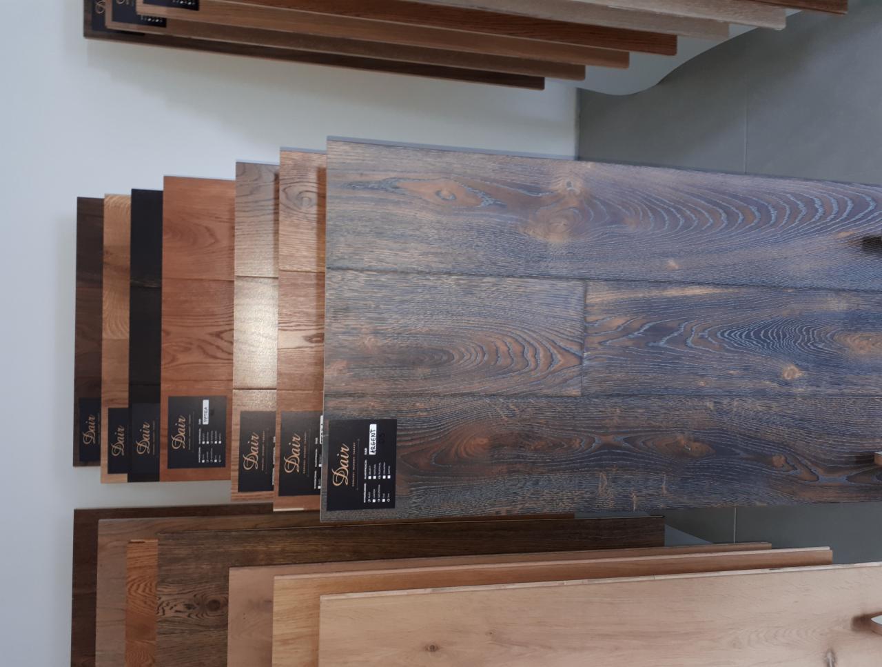 Ekspozycja próbek podłogowych paneli i desek w różnych odcieniach brązu i szarości, z etykietami producenta, ułożonych schodkowo na jasnym tle.