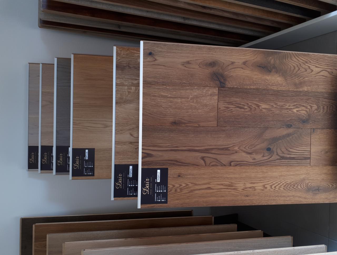 Ekspozycja próbek paneli podłogowych w różnych odcieniach drewna, z widocznymi etykietami producenta Dair, prezentowanych w salonie sprzedaży.