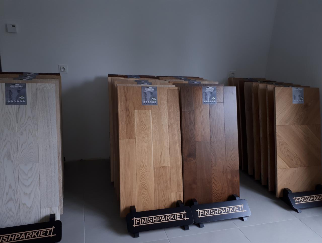 Ekspozycja próbek podłogowych z drewna w różnych odcieniach, umieszczonych na czarnych stojakach z napisem FINISHPARKIET, w jasnym pomieszczeniu.