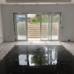 Częściowo ułożona, błyszcząca, czarna podłoga z płytek w jasnym pomieszczeniu, z pomarańczowymi krzyżykami dystansowymi, widok na schody i pojazd budowlany przez przeszklone drzwi przesuwne.