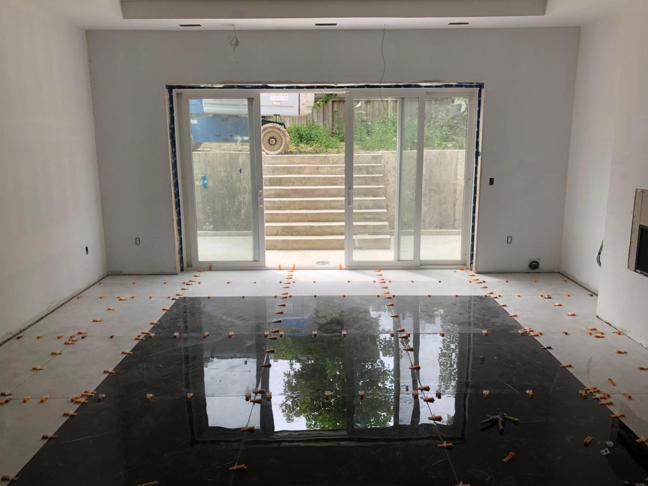 Częściowo ułożona, błyszcząca, czarna podłoga z płytek w jasnym pomieszczeniu, z pomarańczowymi krzyżykami dystansowymi, widok na schody i pojazd budowlany przez przeszklone drzwi przesuwne.