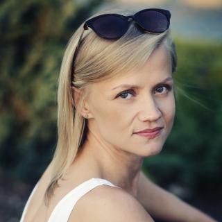 Portret uśmiechniętej kobiety z blond włosami i okularami przeciwsłonecznymi na głowie, spoglądającej przez ramię w kierunku aparatu, na rozmytym zielonym tle.