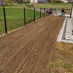 Przygotowanie gleby pod trawnik z rolki: wyrównana ziemia po przekopaniu, widoczny pracownik z grabiami, czarne ogrodzenie siatkowe, budynki w tle.