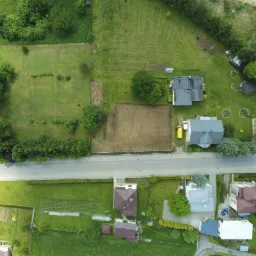 Widok z lotu ptaka na działki z domami, trawnikami i polami uprawnymi, oddzielone drogą i żywopłotem.