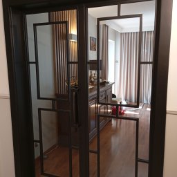 Zakład produkcyjno usługowo hadlowy - Dwuskrzydłowe drzwi wewnętrzne z czarną, metalową ramą i geometrycznymi podziałami szklanymi, odbijające wnętrze salonu z drewnianą komodą i zasłonami.