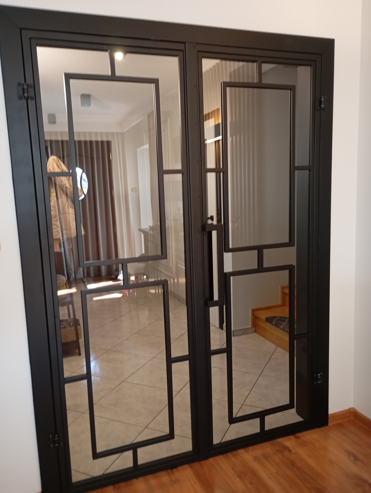 Nowoczesne, dwuskrzydłowe drzwi metalowe z geometrycznymi, prostokątnymi przeszkleniami, czarna rama, widoczne wnętrze domu z parkietem i schodami.